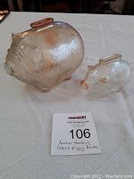 Photo showing both large and small textured glass piggy banks with amber tint, positioned on a white cloth surface with a MaxSold auction card nearby.