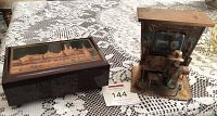 Both music boxes placed on lace tablecloth, showing full view of the copper figurine music box on the right and the wooden box with scenic top on the left.