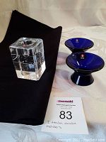 Photo showing 2 blue glass conical Karti candleholders and 1 clear rectangular Arkipelago candleholder on a cloth background with MaxSold tag