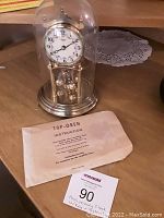 Full view of the Kundo anniversary clock under glass dome with instruction manual.