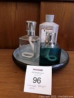 Two Lampe Berger catalytic lamps in square glass bottles, one clear and one teal, along with a bottle of Lampe Berger fragrance and two candle holders on a black tray.