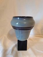 Front view of signed glass vase on black pedestal against a beige background.