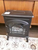Black electric stove-style space heater standing on a patterned carpet with a wooden cabinet in the background.