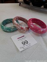 Three mother of pearl mosaic bangle bracelets in green, light pink, and reddish pink, displayed on white surface with seller label.
