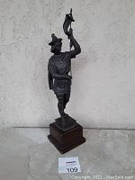 Full view of the dark spelter figurine depicting a standing warrior holding a banner on wooden base against a plain textured wall.