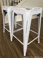 Side and angled view of two distressed white metal bar stools standing on wood floor near staircase. Shows tapered legs and crossbars.