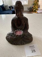 Front view of a metal Buddha statue holding a flower-shaped tea light candle holder with a pink candle inside on a white countertop in a bright room.