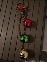 Photo of three large metallic jingle bells in red, green, and gold on a twisted red and white cord with holly and poinsettia decoration