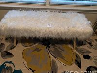 Front view of the ecru faux fur bench showing long shaggy faux fur upholstery and four light wooden legs on a multi-colored rug beneath a window.