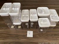 Nine clear plastic food storage containers with white lids arranged on wooden floor, showing varying sizes and shapes including rectangular and square containers.