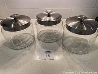 Three clear glass jars with stainless steel lids, placed on a kitchen counter with white tiled backsplash.