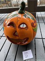 Front view of orange ceramic pumpkin with carved face and green stem on top, showing painted leaf and vine accents and hollow facial cutouts.