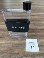 Front view of the clear HYDRTE flask with black grip and cap on wooden surface with lot number card.