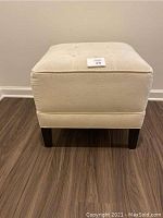 Front view of the square ecru ottoman showing button tufting and dark wooden legs.