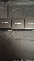 Three closed black boxes labeled 'Smart Watch' on a black surface, indicating the lot includes three smart watches in packaging.