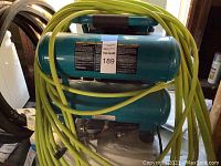 Full view of teal Makita dual-stack pancake air compressor with coiled yellow-green hose draped over top