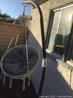 Overall view of outdoor hanging swing chair with thick round gray cushion and macrame netting, attached on black curved metal stand, placed outside near a house wall and window.