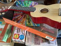 Red ukulele on top of various craft boxes and game packages, showing part of the lot's musical instrument and craft items.