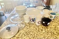 Wide shot showing collection of white plates, coffee mugs, large white bowl, multiple smaller bowls, clear glass lid with a black knob, and three bowl tops on a granite countertop.