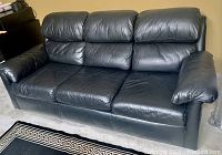 Black leather couch with three cushions and wide padded armrests, showing normal wear, on light floor.