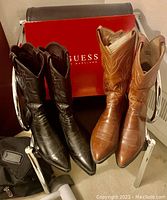 Pair of Guess ladies boots in dark brown and pair of Tony Lama brown men's boots on chair with Guess box behind.