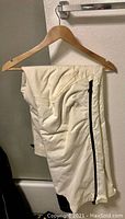 White men's ski pants hanging on hanger, showing zipper detail on side and ankle adjustment straps.
