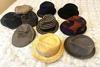 Top view showing all 10 men's hats laid out on a white textured surface, including fedoras, trilbys, flat caps, and a straw fedora.