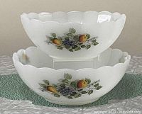 Two white milk glass bowls stacked, both have a fruit design including peaches and grapes, scalloped edges visible