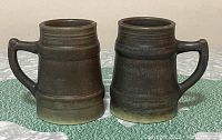 Two large pottery mugs with dark brown to greenish glaze, side by side showing front and side profiles.