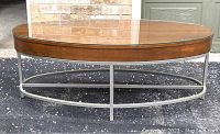 Full view of oval designer coffee table showing wood and glass top with iron base on pavement in front of brick wall and door.
