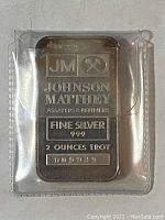 Silver bar front face showing Johnson Matthey branding, silver purity (.999), weight (2 ounces troy), and serial number 009939 in a plastic protector.