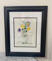 Full view of framed print showing black frame and white mat with colorful flowers and hands drawing.