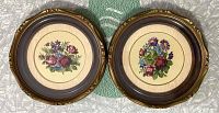 Two matching vintage circular framed needlepoint floral artworks laying flat on packaging material. Ornate faux wood frames in brown with gold accents, frames measure approximately 10 3/4 inches in diameter.