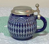 Left side view of cobalt blue German stein with decorative diamond and scroll patterns and pewter lid with rounded knob.