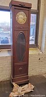 Front full view of the grandfather clock near window showing overall condition, wooden cabinet design, and clock face.
