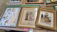 Three framed pieces on table including the colorful embroidery with broken glass and two vintage illustration prints in different frames.