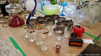 Photo showing multiple items including a clear glass swan sculpture, red glass apple, green glass pear, the Avon cologne bottle, a set of drinking glasses with gold rims, and a footed glass bowl with green stem and white bowl.
