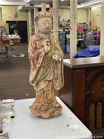 Full front view of the Asian sculpture showing standing figure with weathered reddish paint and chipped surface, including missing left index finger.