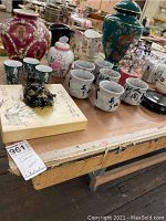 Photo showing large ginger jars, ceramic mugs with Asian characters, small ginger jars, a black lacquer dish, and a decorative piece on a wooden table.