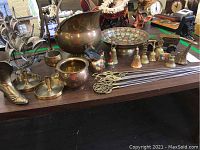 Top-down view of the brass items on a table showing multiple brass collectibles including a large pitcher, decorative bowl, model ship, shoe, bells, and rods