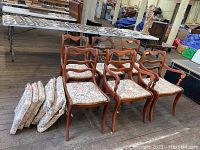 Overall view of 6 wooden dining chairs with floral patterned seat cushions and 6 additional matching cushions stacked beside them.