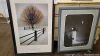 Framed art print "Solitude" by David Lorenz Winston with a lone tree and fencing in a snowy field with a black frame and white mat next to a framed black-and-white photo of the Hale Bop comet over a lighthouse.