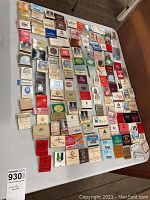 Top view of approximately 80 assorted matchbooks from hotels, restaurants, and resorts laid out on a white sheet on the floor.