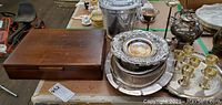 Overview of wooden flatware case, silver and glass serving pieces including hammered ice bucket with lid, round serving tray, 6 cordial cups, teapot, and wine glass
