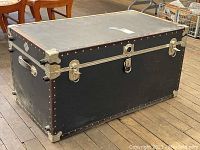 Front view of Ashworth black foot locker trunk showing front lock, horizontal metal bands, rivets, and handles on sides.