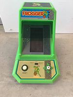 Front view of official Frogger electronic game showing green casing, screen with Frogger artwork, joystick, reset button, and skill switch.