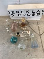 Nine assorted mostly empty perfume bottles on ground with white box behind