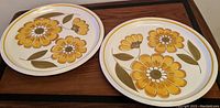 Two round ceramic platters with yellow and brown floral Happy design lying side by side on a wooden surface.