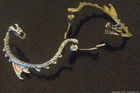 Close-up photo showing a silver dragon ear cuff and a gold dragon ear cuff, each with detailed scales, dragon head, and wings, designed to wrap around the ear.