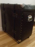 Front and side view of black vintage drum/trap case with metal handles, riveted construction, metal corner protectors, secured with two exterior straps. Shows wear consistent with vintage condition.